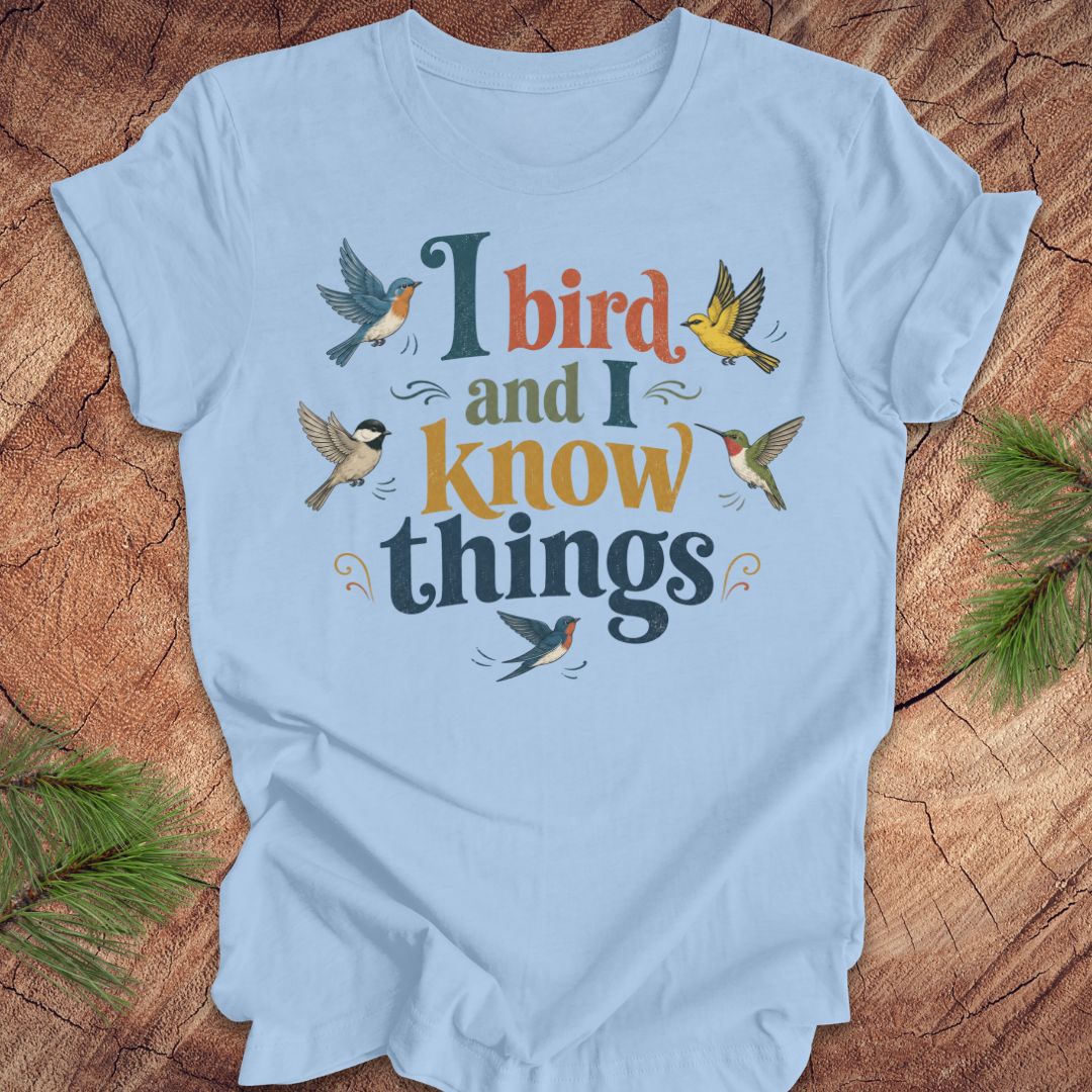 A light blue short-sleeve t-shirt with a graphic print reading 'I bird and I know things' in stylized dark teal and mustard lettering, surrounded by illustrated birds, laid flat on a rustic wood surface with pine branches at the corners.