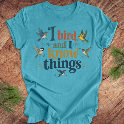 A turquoise blue short-sleeve t-shirt with a graphic print that reads 'I bird and I know things' in dark blue and orange lettering, surrounded by illustrated birds, laid flat on a rustic wood background with pine branches in the corners.