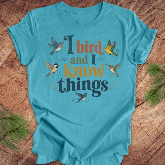 A turquoise blue short-sleeve t-shirt with a graphic print that reads 'I bird and I know things' in dark blue and orange lettering, surrounded by illustrated birds, laid flat on a rustic wood background with pine branches in the corners.