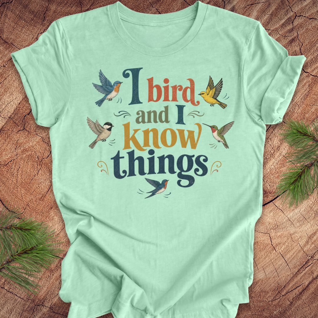 A mint green short-sleeve t-shirt with a colorful graphic print that reads 'I bird and I know things' surrounded by illustrated birds, laid flat on a rustic wood background with pine branches in the corners.