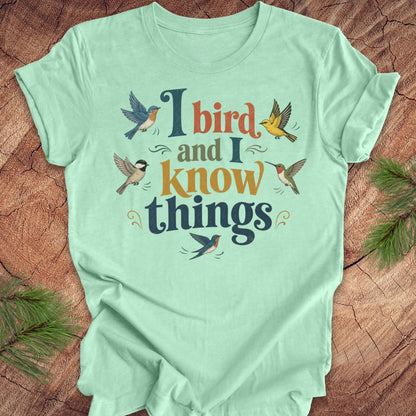A mint green short-sleeve t-shirt with a colorful graphic print that reads 'I bird and I know things' surrounded by illustrated birds, laid flat on a rustic wood background with pine branches in the corners.