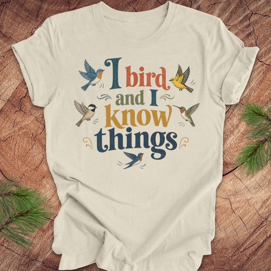 A beige short-sleeve t-shirt with a colorful graphic print reading 'I bird and I know things' surrounded by illustrated birds, laid flat on a rustic wood background with pine branches in the corners.