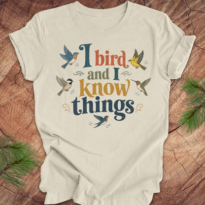 A beige short-sleeve t-shirt with a colorful graphic print reading 'I bird and I know things' surrounded by illustrated birds, laid flat on a rustic wood background with pine branches in the corners.