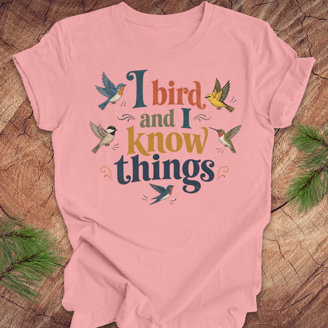 A light pink short-sleeve t-shirt with a colorful graphic that reads 'I bird and I know things' surrounded by small birds, laid flat on a rustic wood background with pine branches in the corners.