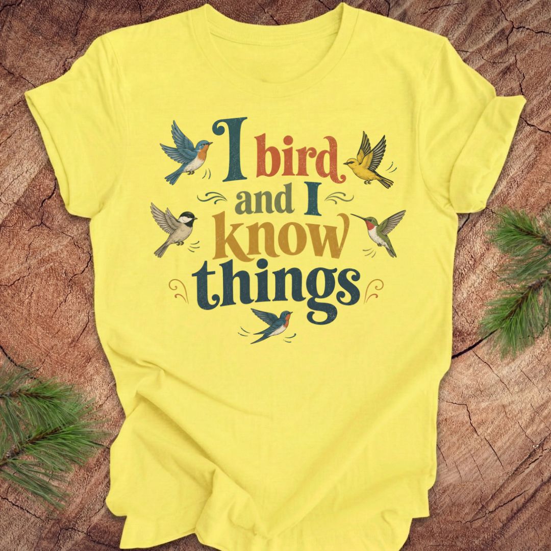 A bright yellow short-sleeve t-shirt laid flat on a rustic wood surface, featuring a colorful graphic print that reads 'I bird and I know things' surrounded by illustrated birds. Green pine branches are placed in the corners as props.