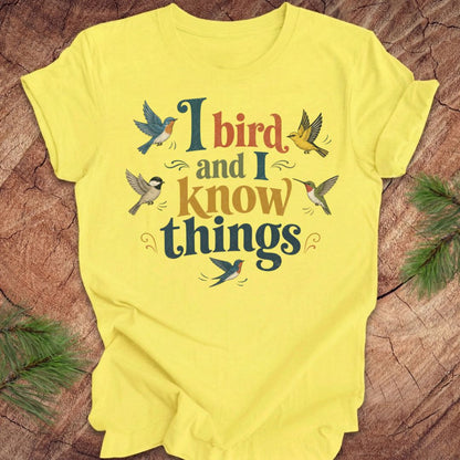 A bright yellow short-sleeve t-shirt laid flat on a rustic wood surface, featuring a colorful graphic print that reads 'I bird and I know things' surrounded by illustrated birds. Green pine branches are placed in the corners as props.