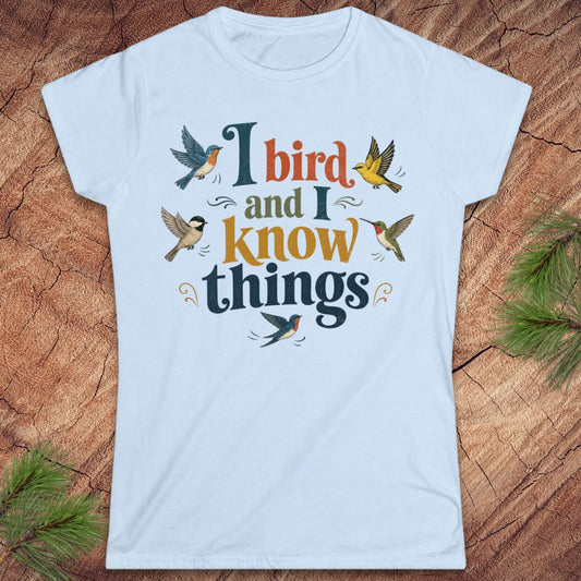 A light blue crewneck t-shirt featuring a colorful graphic print that reads 'I bird and I know things' surrounded by illustrated birds, laid flat on a rustic wood background with pine needles.