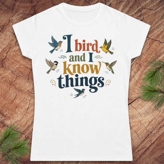 A white short-sleeve crewneck t-shirt with a colorful graphic print that reads 'I bird and I know things' surrounded by illustrated birds, laid flat on a rustic wood background with pine needles in the corners.