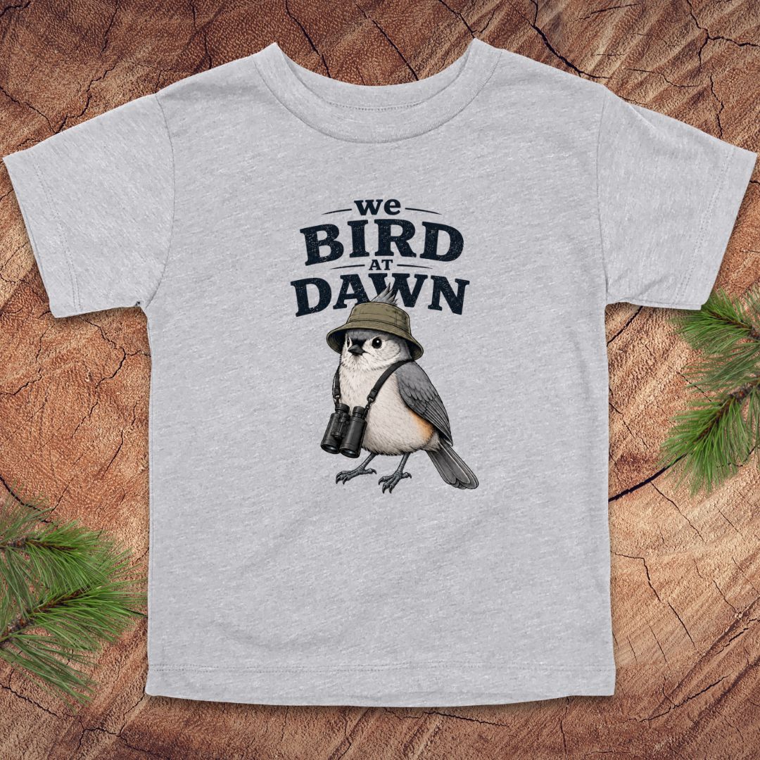 Gray t-shirt with a bird graphic and text 'we BIRD at DAWN' on a wooden surface.