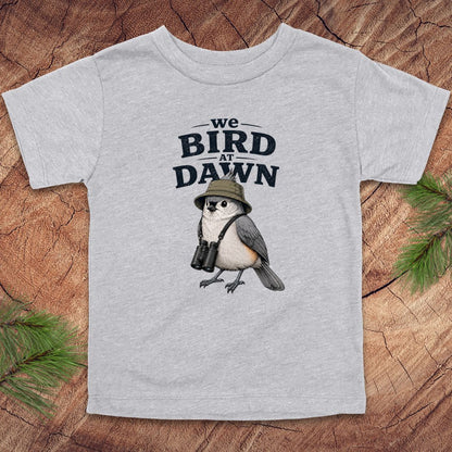 Gray t-shirt with a bird graphic and text 'we BIRD at DAWN' on a wooden surface.