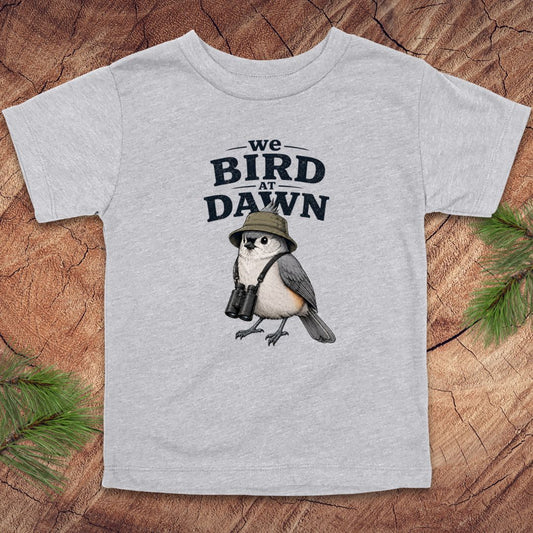 Gray t-shirt with a bird graphic and text 'we BIRD at DAWN' on a wooden surface.