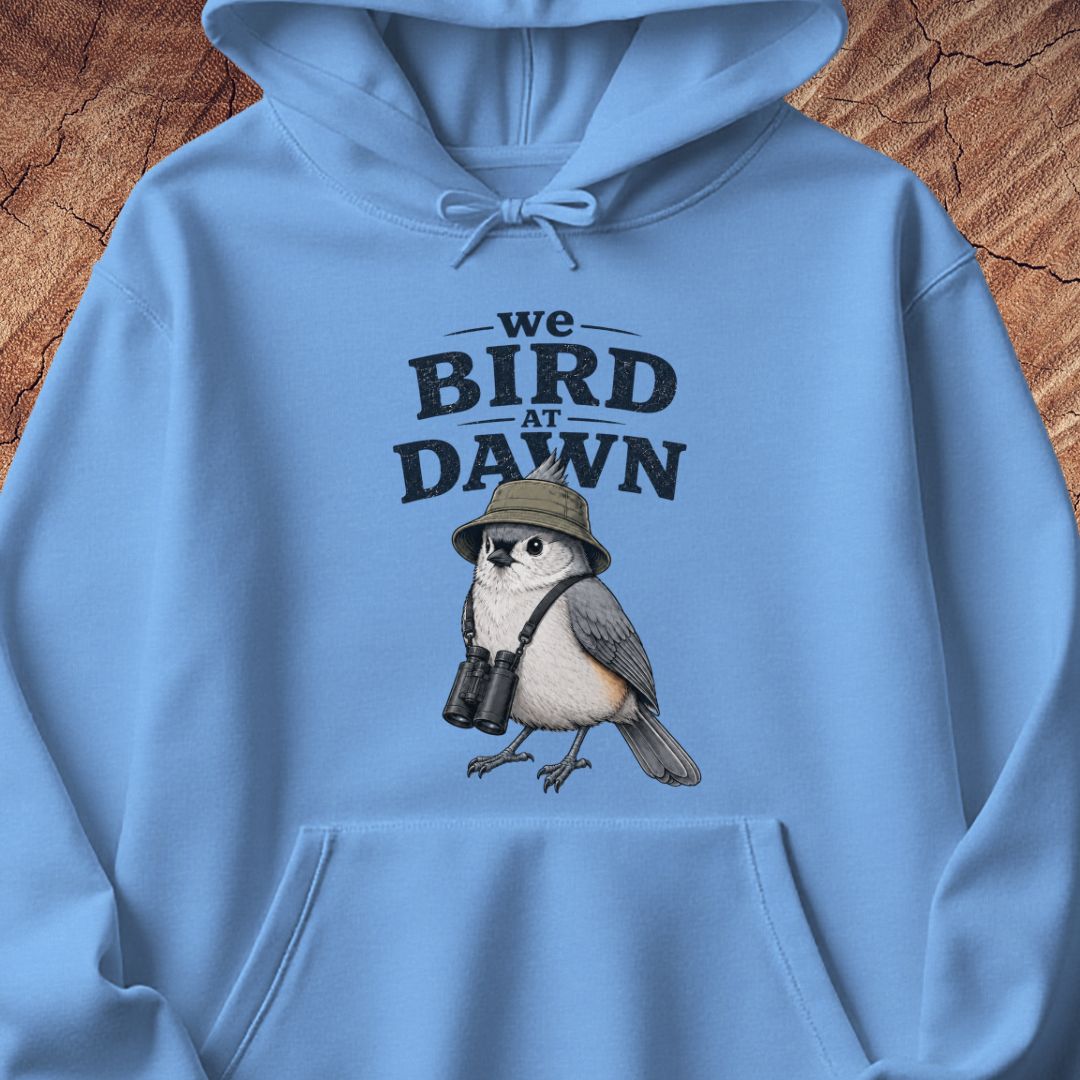 A light blue hoodie laid flat on a rustic wood background, featuring a front graphic that reads 'we BIRD AT DAWN' above an illustrated bird wearing a safari hat and binoculars.