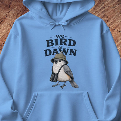 A light blue hoodie laid flat on a rustic wood background, featuring a front graphic that reads 'we BIRD AT DAWN' above an illustrated bird wearing a safari hat and binoculars.