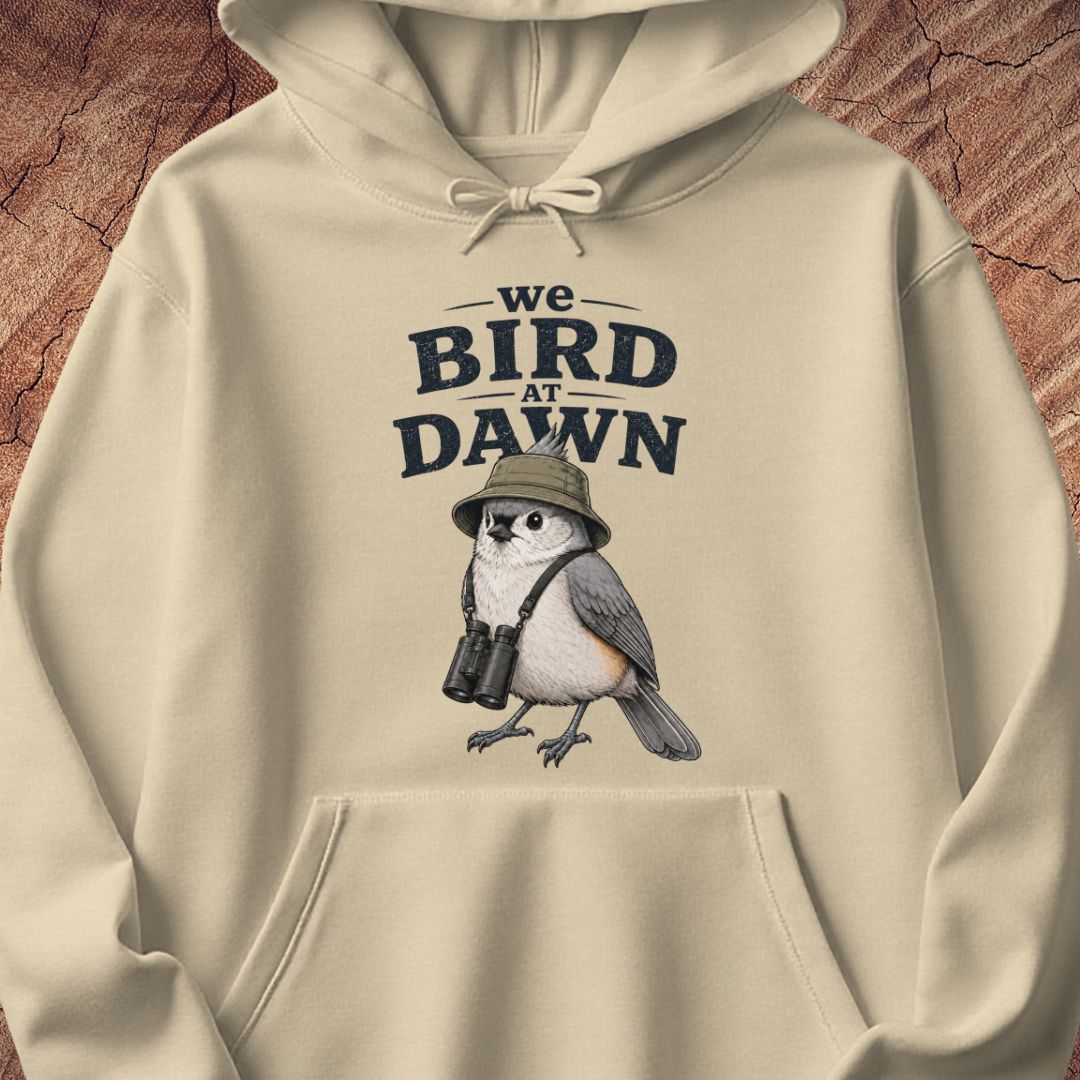 A cream-colored hoodie featuring a graphic print of a bird wearing a hat and binoculars under the text 'we BIRD AT DAWN'. The hoodie is displayed flat against a textured brown background resembling tree bark.