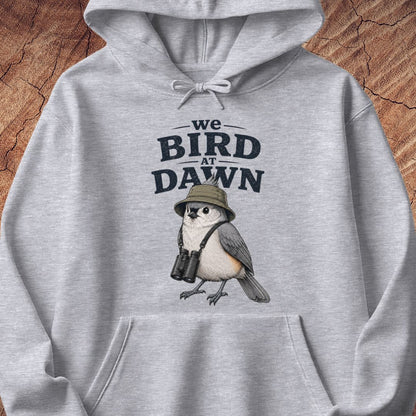 A light gray heathered hoodie laid flat on a textured wood background, featuring a printed graphic of a bird wearing a hat and binoculars beneath the text 'we BIRD AT DAWN'.