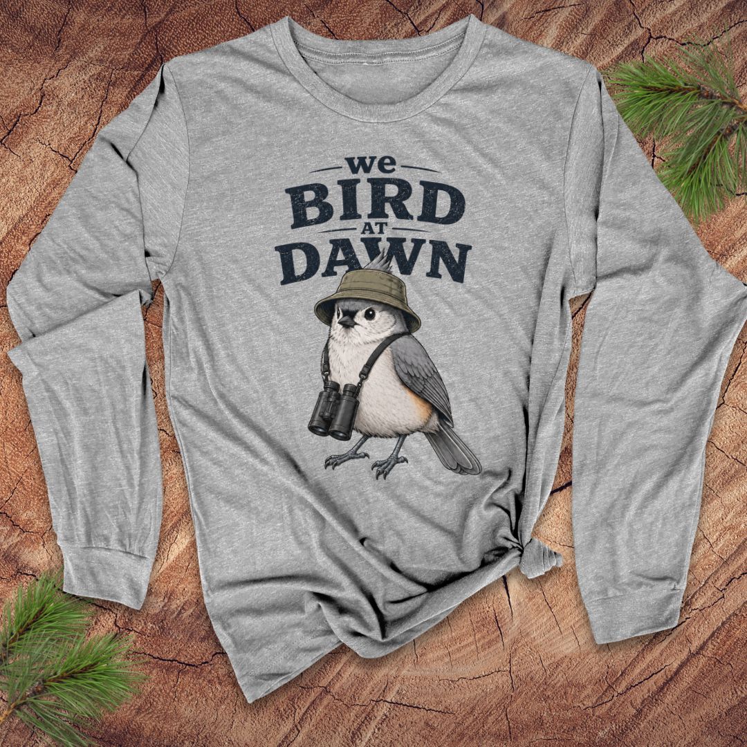 Gray long-sleeve tshirt with a titmouse and text 'we bird at dawn' on a wood background.