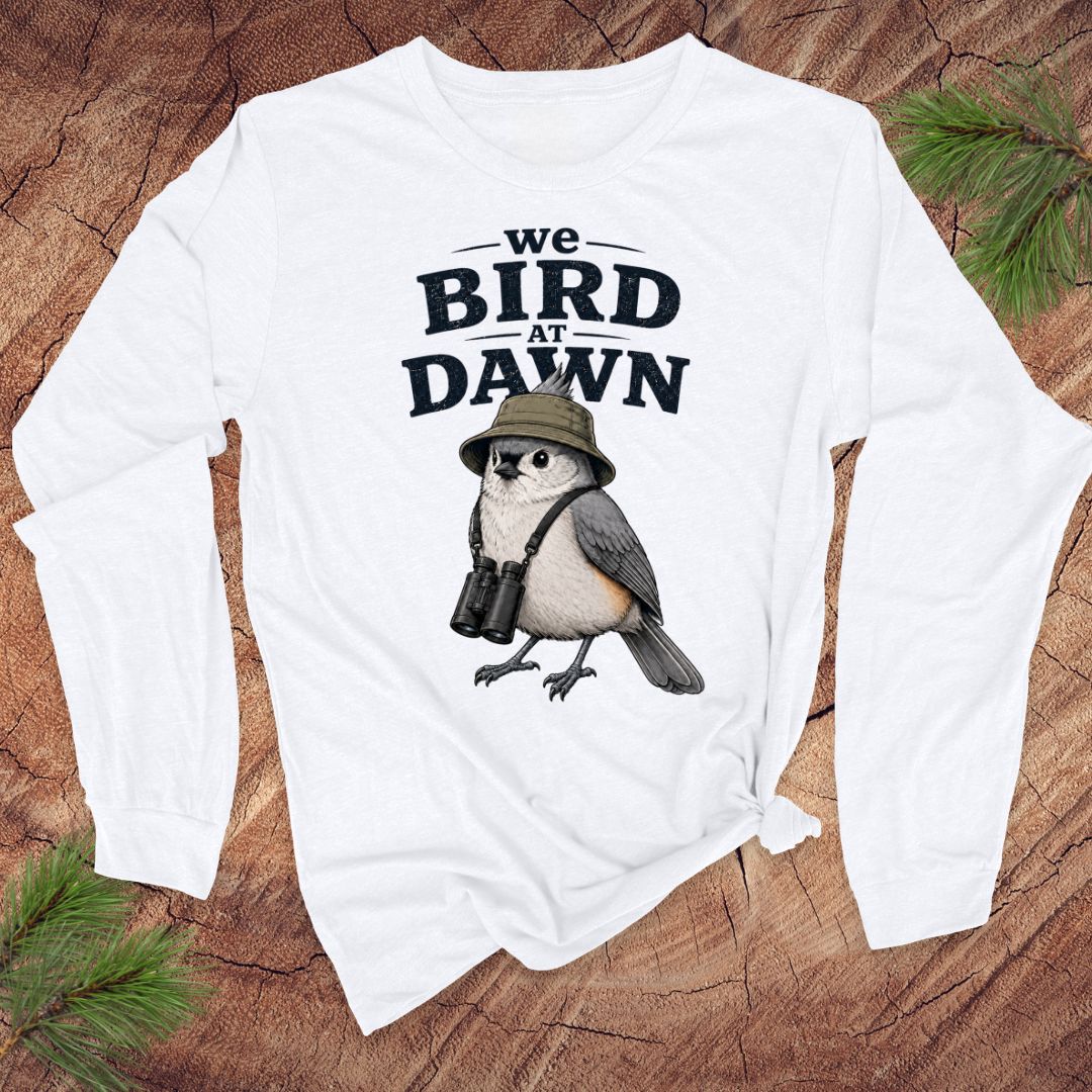 White long-sleeve tshirt with a titmouse and text 'we bird at dawn' on a wood background.