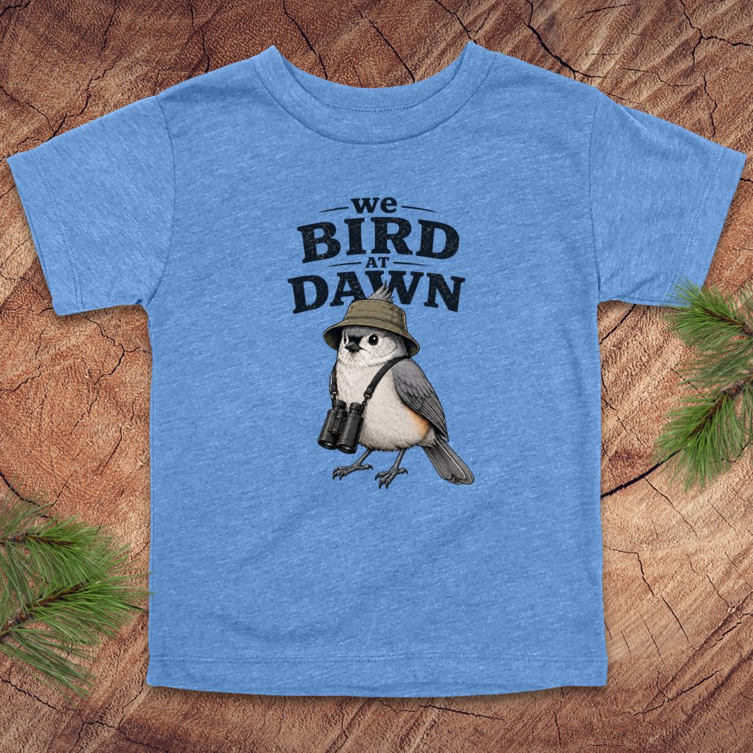 Blue t-shirt with a bird graphic and text 'we BIRD at DAWN' on a wooden surface.