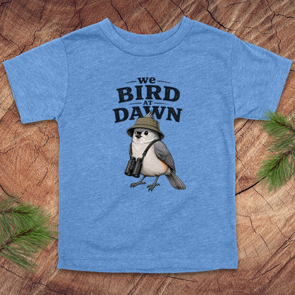 Blue t-shirt with a bird graphic and text 'we BIRD at DAWN' on a wooden surface.