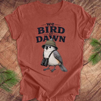 Rust colored t-shirt with a titmouse graphic and text 'we BIRD at DAWN' on a wooden surface.