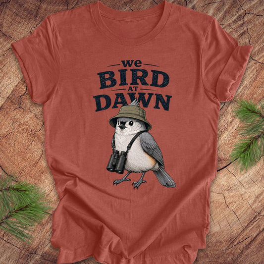Rust colored t-shirt with a titmouse graphic and text 'we BIRD at DAWN' on a wooden surface.