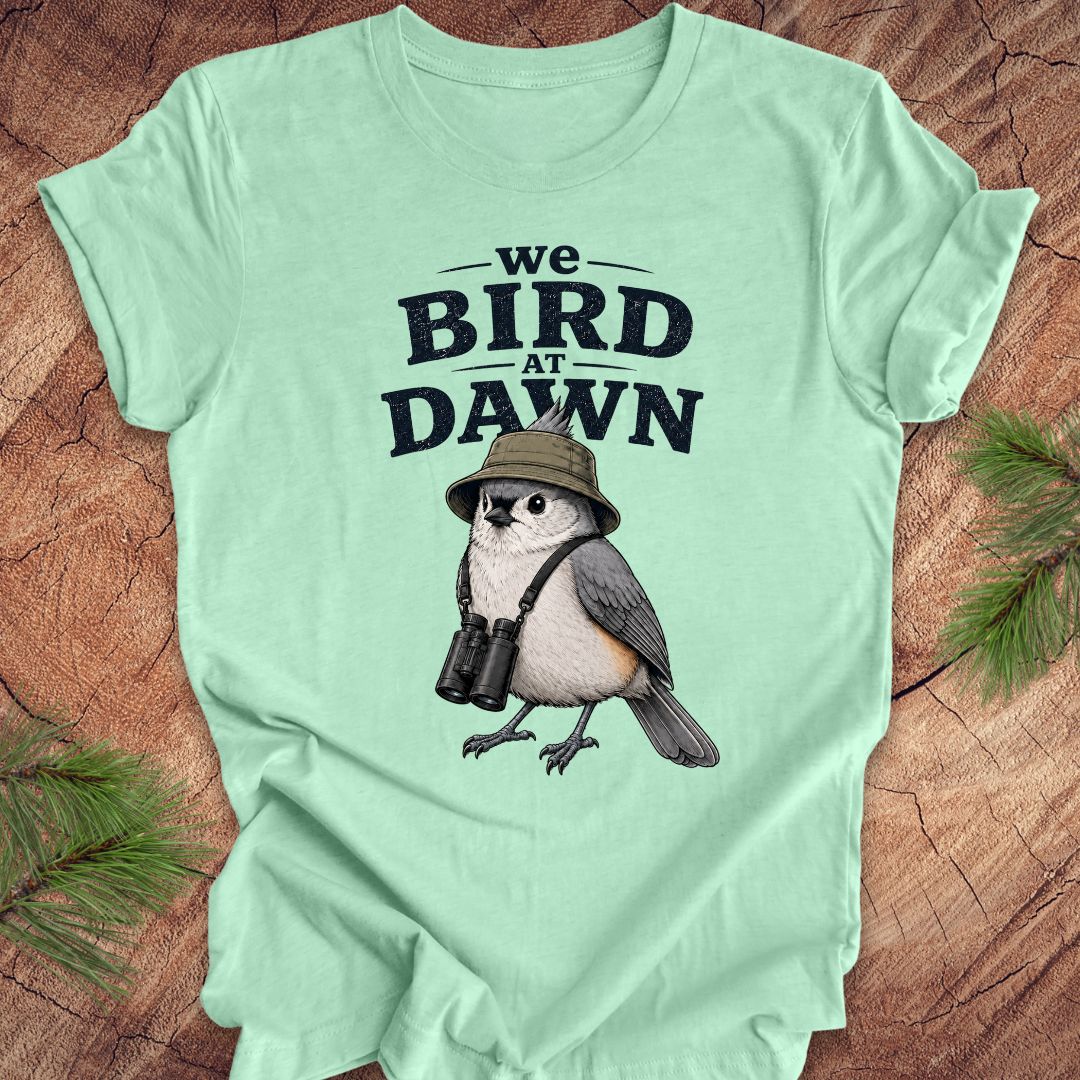 Green t-shirt with a titmouse graphic and text 'we BIRD at DAWN' on a wooden surface.