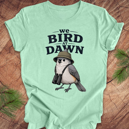 Green t-shirt with a titmouse graphic and text 'we BIRD at DAWN' on a wooden surface.