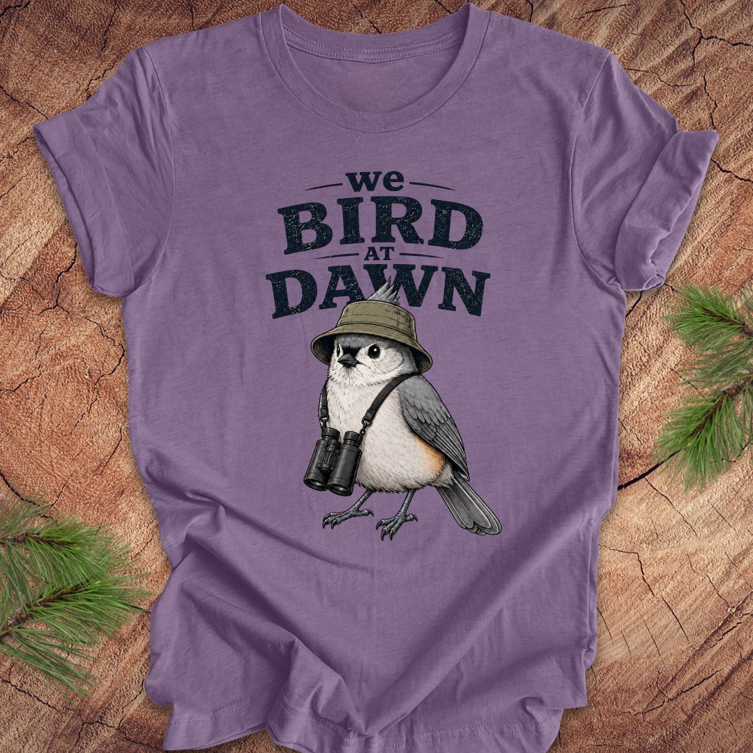 Purple t-shirt with a titmouse graphic and text 'we BIRD at DAWN' on a wooden surface.
