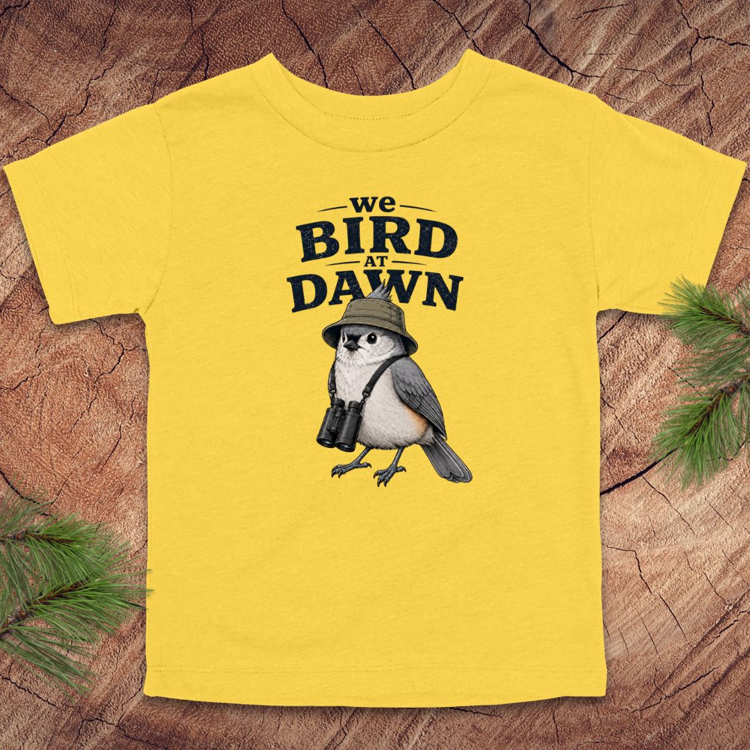 Yellow t-shirt with a bird graphic and text 'we BIRD at DAWN' on a wooden background.