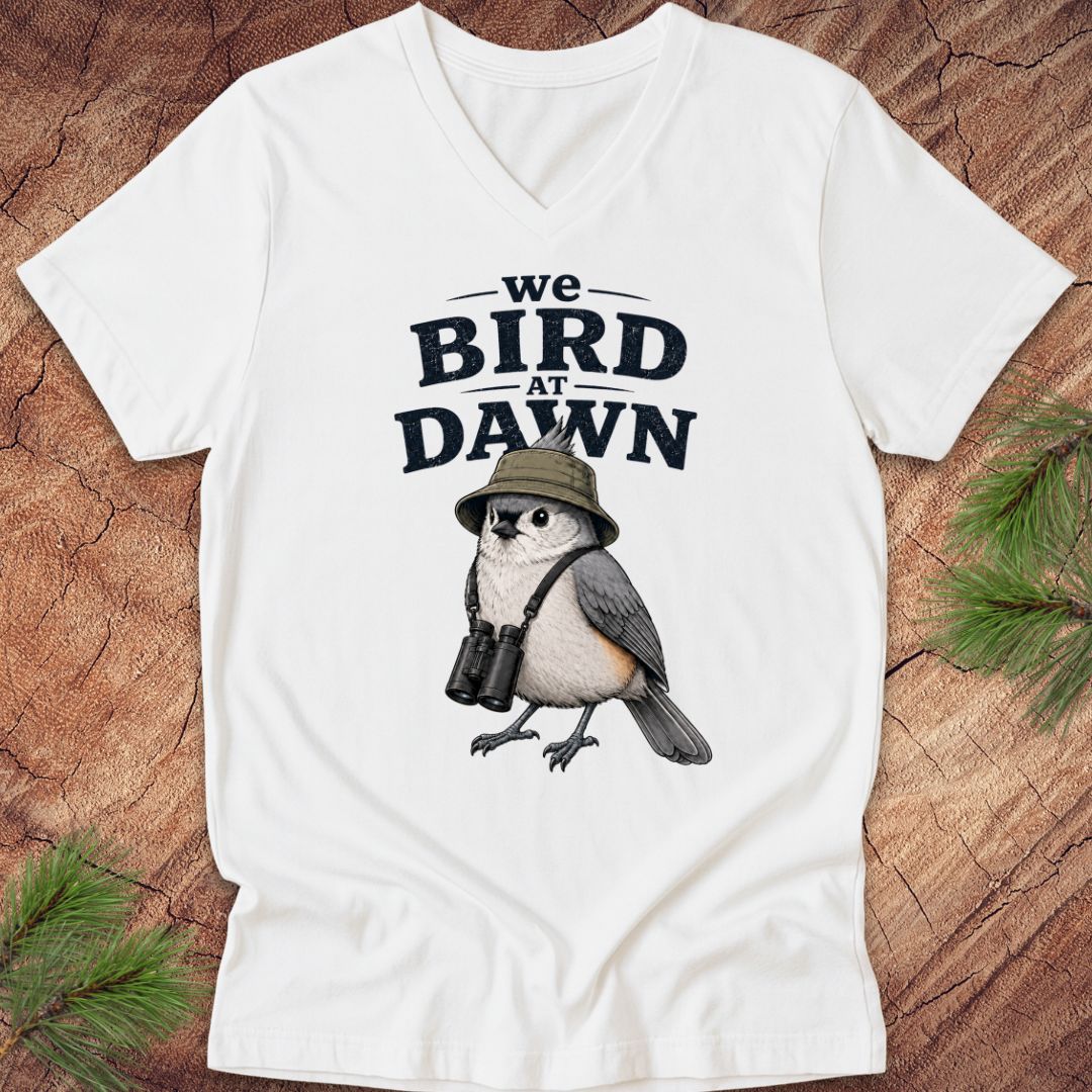 White t-shirt with a bird graphic and text 'we BIRD at DAWN' on a wooden surface.
