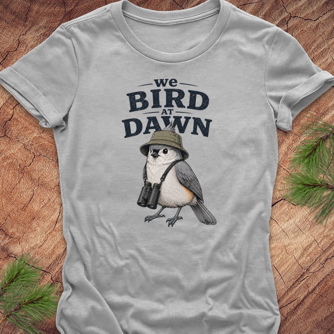 A light gray short-sleeve t-shirt laid flat on a rustic wood background with pine branches in the corners. The shirt features a graphic print of a bird wearing a safari hat and binoculars, with the text 'we BIRD AT DAWN' above it.