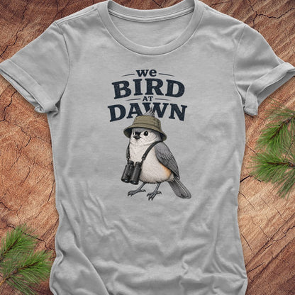 A light gray short-sleeve t-shirt laid flat on a rustic wood background with pine branches in the corners. The shirt features a graphic print of a bird wearing a safari hat and binoculars, with the text 'we BIRD AT DAWN' above it.