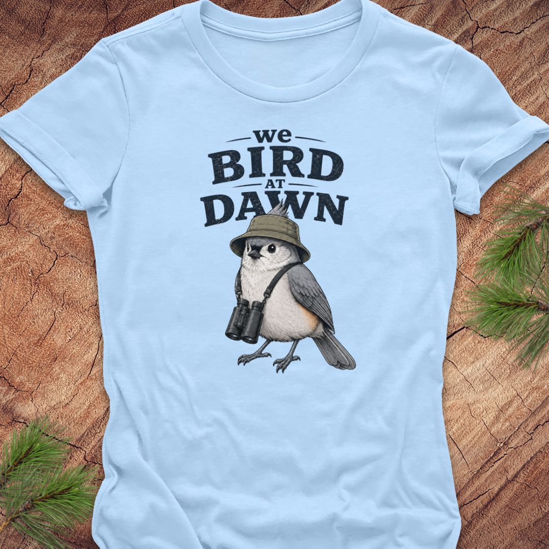 A light blue short-sleeve t-shirt laid flat on a rustic wood surface with pine branches in the corners. The shirt features a graphic print of a bird wearing a safari hat and binoculars beneath the text 'we BIRD AT DAWN' in black.