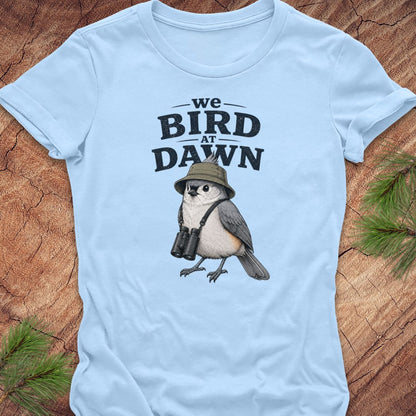 A light blue short-sleeve t-shirt laid flat on a rustic wood surface with pine branches in the corners. The shirt features a graphic print of a bird wearing a safari hat and binoculars beneath the text 'we BIRD AT DAWN' in black.