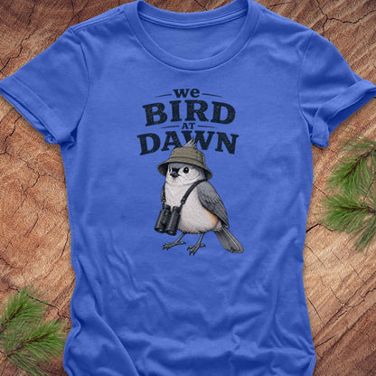 A royal blue short-sleeve t-shirt laid flat on a rustic wood surface, featuring a printed graphic of a bird wearing a safari hat and binoculars with the text 'we BIRD AT DAWN'. Green pine branches are placed around the edges as styling props.