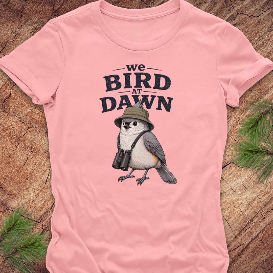 A light pink short-sleeve t-shirt laid flat on a textured wood background with pine branches in the corners. The shirt features a graphic print of a bird wearing a safari hat and binoculars, with the text 'we BIRD AT DAWN' above it.