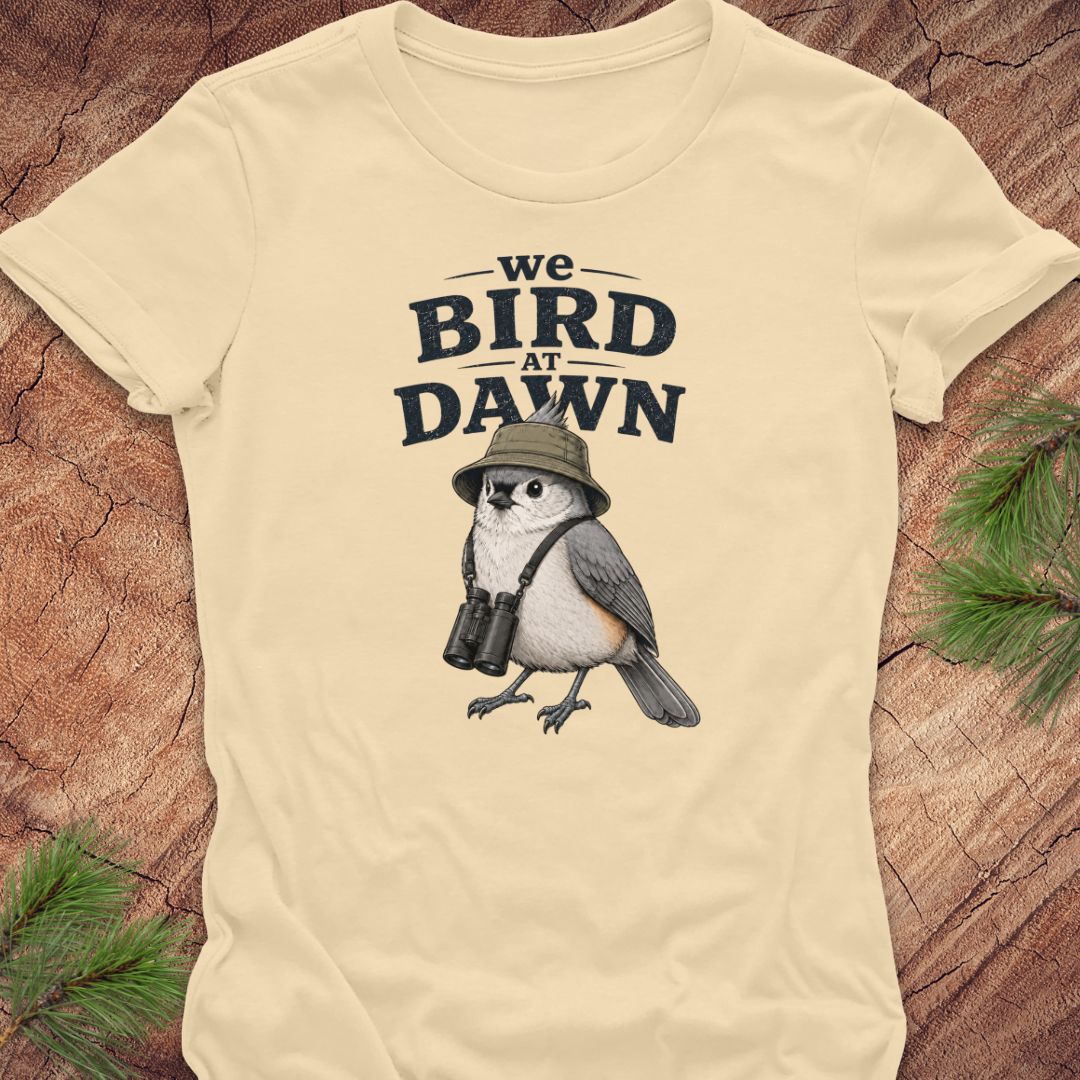 A cream-colored short-sleeve t-shirt with a graphic print of an owl wearing a safari hat and binoculars under the text 'we BIRD AT DAWN'. The shirt is laid flat on a textured wood background with pine branches in the corners.
