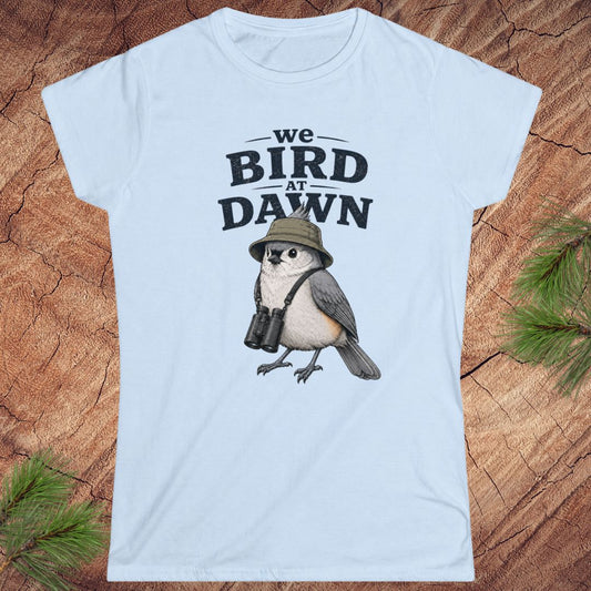 Light blue t-shirt with a bird graphic and text 'we BIRD at DAWN' on a wooden surface.