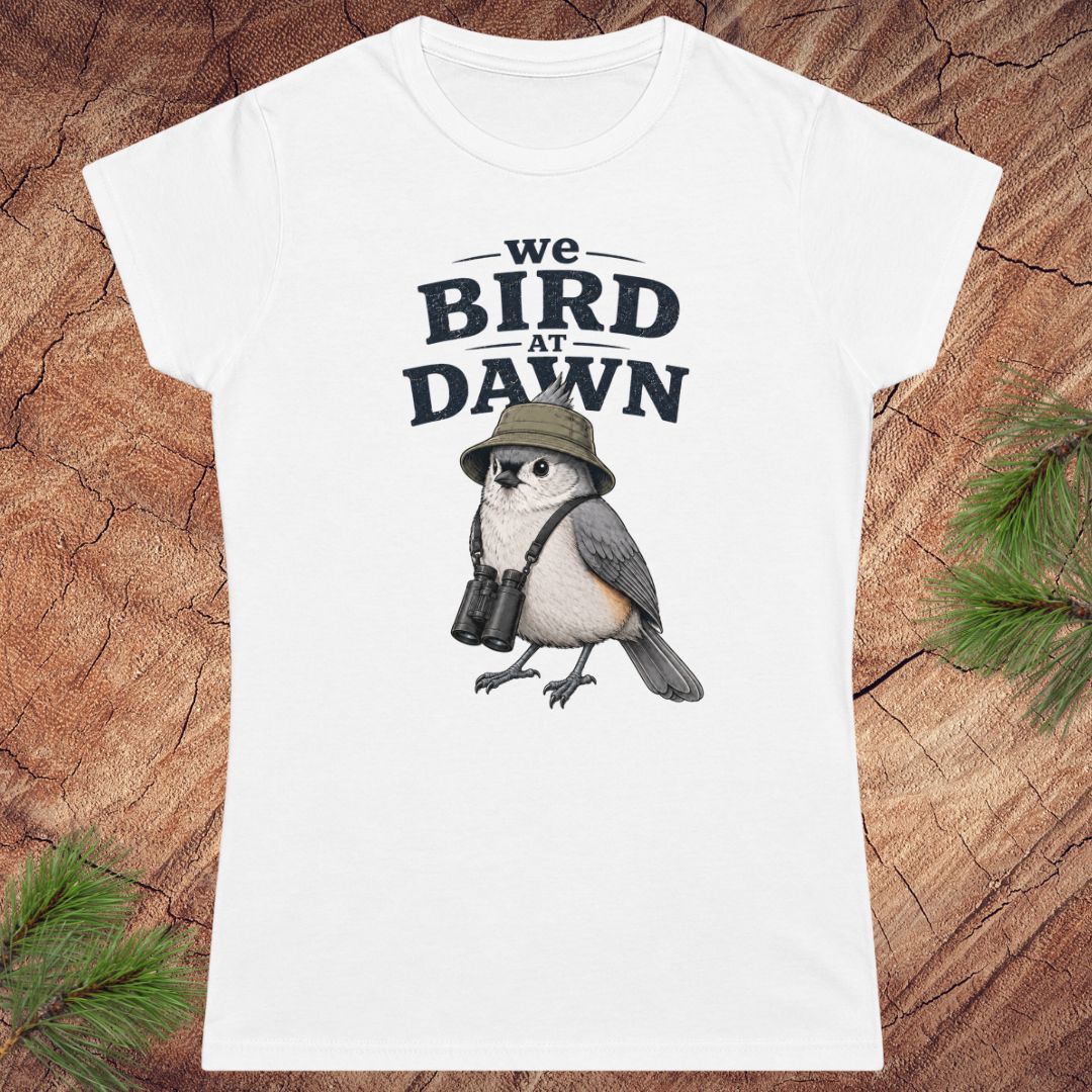 White t-shirt with a bird graphic and text 'we BIRD at DAWN' on a wooden surface.