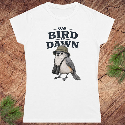 White t-shirt with a bird graphic and text 'we BIRD at DAWN' on a wooden surface.