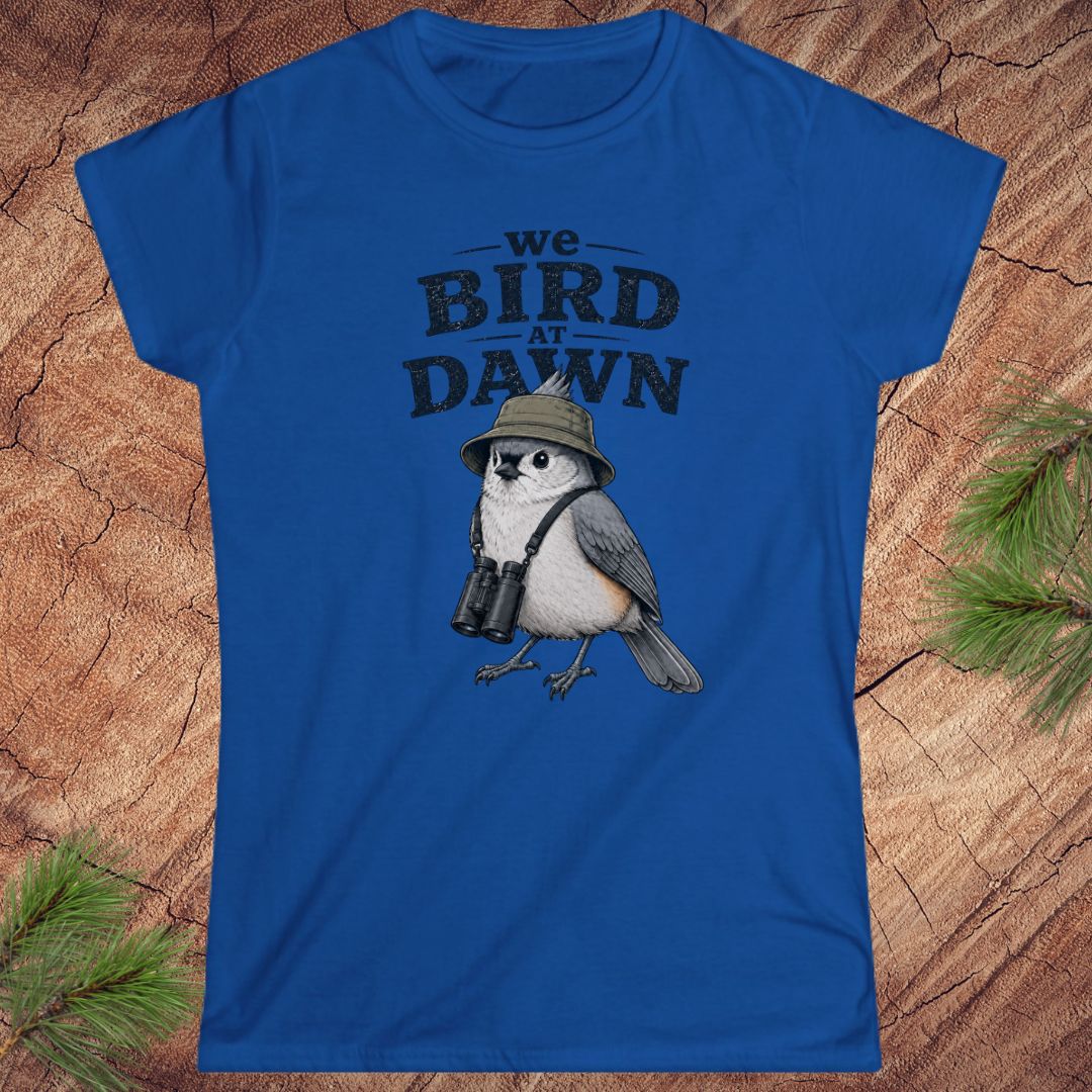Blue t-shirt with a bird graphic and text 'We Bird at Dawn' on a wooden surface.