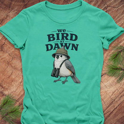 A mint green short-sleeve t-shirt laid flat on a rustic wood background with pine branches in the corners. The shirt features a graphic print of a bird wearing a safari hat and binoculars beneath the text 'we BIRD AT DAWN'.