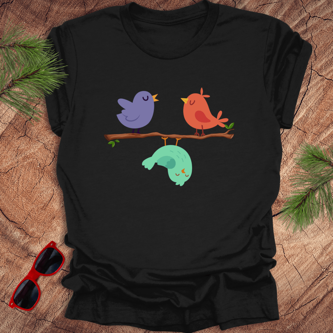 Black t-shirt with colorful birds design on a wooden surface with sunglasses and pine branches.