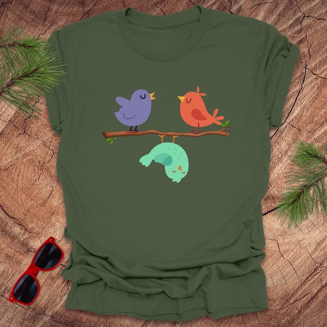 Green t-shirt with cartoon birds design on a wooden surface with sunglasses and pine branches.