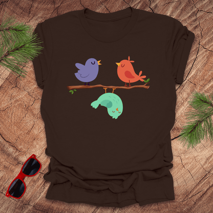 Brown t-shirt with colorful birds design on a wooden surface with sunglasses and pine branches.