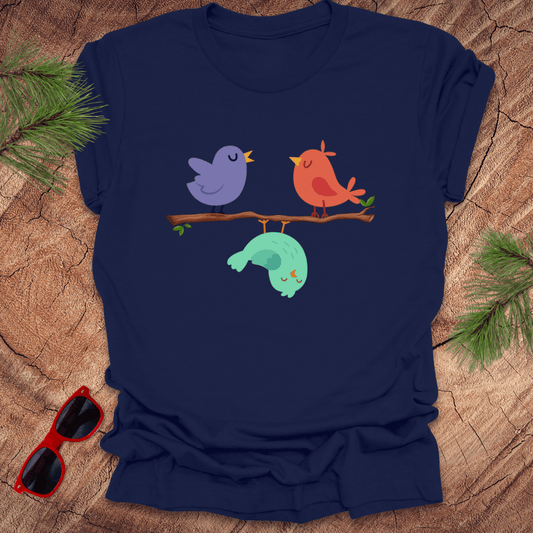 Navy blue t-shirt with cartoon birds and caterpillar design on a wooden surface with sunglasses and pine branches.