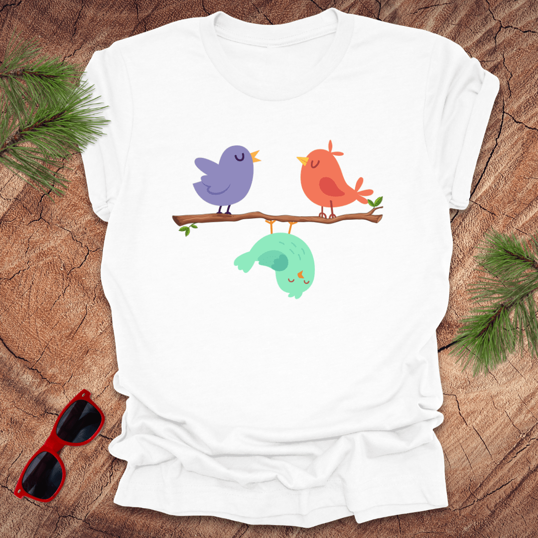 White t-shirt with colorful birds design on a wooden surface with sunglasses and greenery.