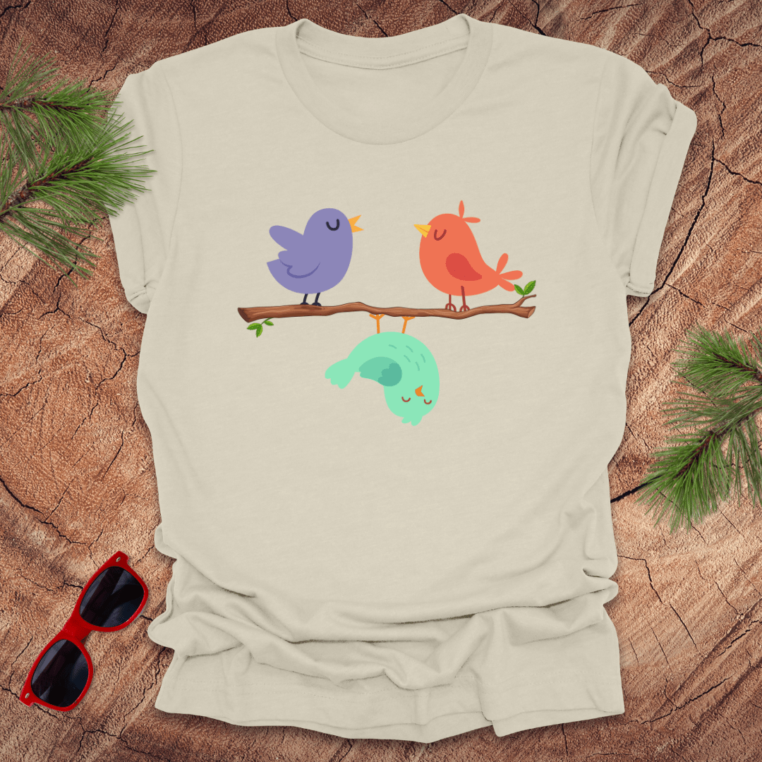Beige t-shirt with cartoon birds on a branch on a wooden surface with sunglasses and greenery.