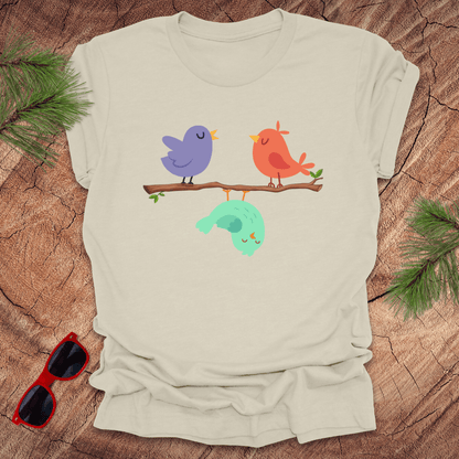Beige t-shirt with cartoon birds on a branch on a wooden surface with sunglasses and greenery.