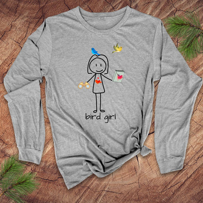 Grey long-sleeve shirt with 'bird girl' text and stick figure girl with binoculars and guide book, on a wooden surface with pine branches.